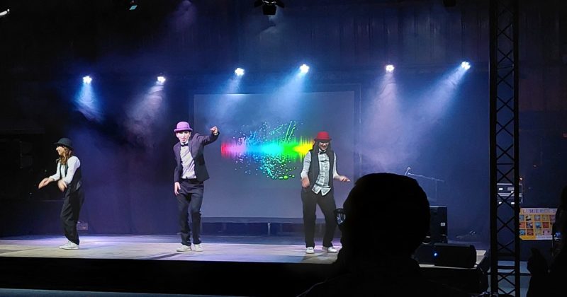 Trois artistes en spectacle sur scène, avec éclairage dynamique et effets visuels en arrière-plan.