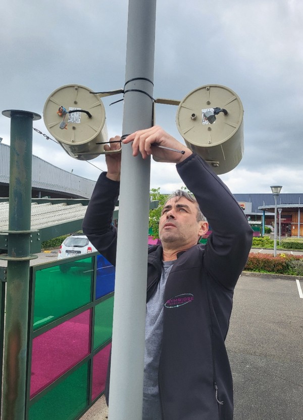 Technicien installant du matériel sonore sur un poteau dans un espace extérieur, sous un ciel nuageux.