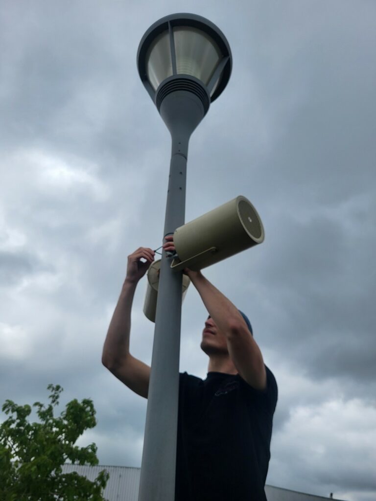 Technicien installant des haut-parleurs sur un lampadaire sous un ciel nuageux.