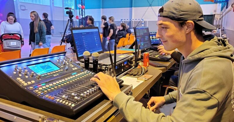Technicien réglant une console de son lors d'un événement, avec équipements audio et public en arrière-plan.
