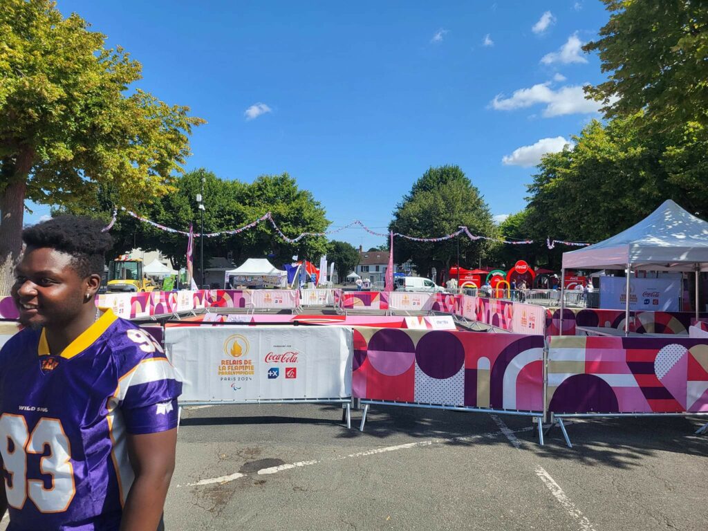 Installation d'un événement extérieur avec des barrières décoratives et un participant en tenue sportive. Ciel bleu et stands animés en arrière-plan.