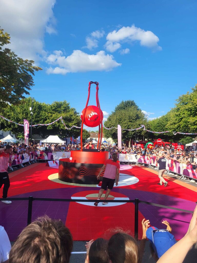 Événement festif en plein air avec des participants autour d'une grande installation rouge au sol, sous un ciel ensoleillé.