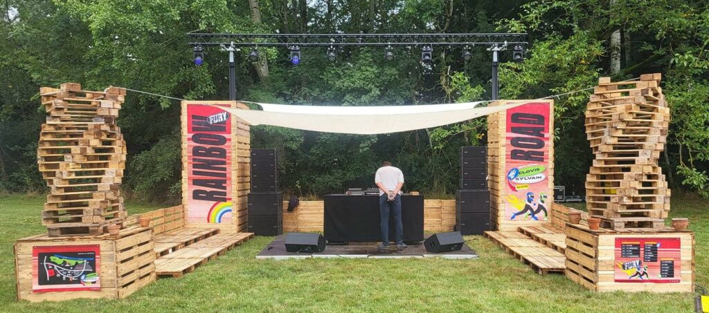 Scène de festival avec matériel audio, éclairage et décor en palettes de bois. Un DJ se prépare à jouer dans un environnement verdoyant.