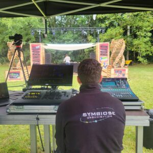 Technicien de son et lumière de Symbiose Organisation préparant un événement en extérieur, avec équipement audio et écran de contrôle.