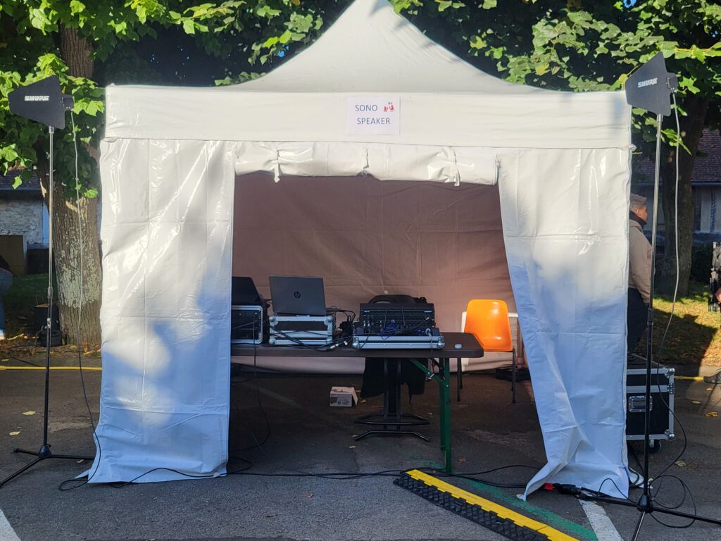 Tente pour la location de matériel de sono, avec tables, ordinateurs et systèmes audio sous un ciel ensoleillé.