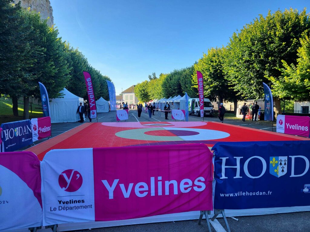 Vue d'un espace d'événement avec des stands, des banderoles Yvelines et une zone marquée au sol, entouré d'arbres verts.