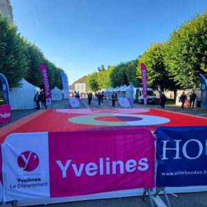 Vue d'un espace d'événement avec des stands, des banderoles Yvelines et une zone marquée au sol, entouré d'arbres verts.