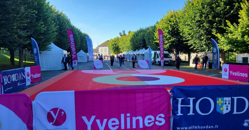 Vue d'un espace d'événement avec des stands, des banderoles Yvelines et une zone marquée au sol, entouré d'arbres verts.