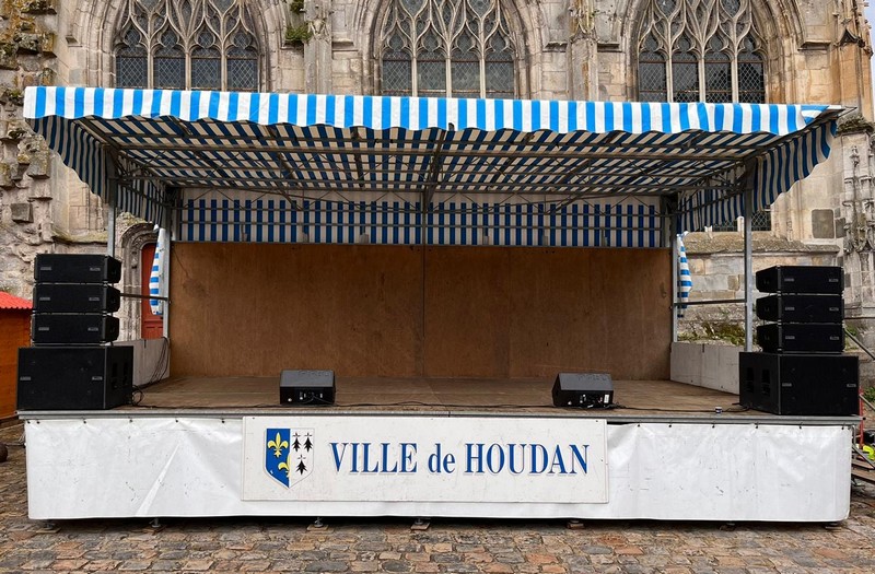 Scène de spectacle en plein air avec sonorisation, marquée "VILLE de HOUDAN", sous un chapiteau à rayures bleues et blanches.